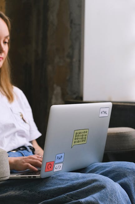 A person's hands are positioned over a sleek, silver laptop displaying a dark-themed coding interface with colorful lines of programming code on the screen. The background is softly blurred, with a hi